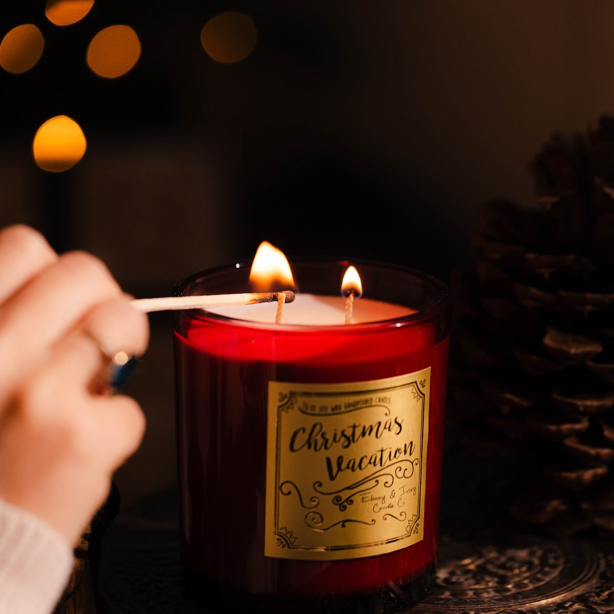 Christmas soy Candle being lit with a pine cone and blurred lights in the background.