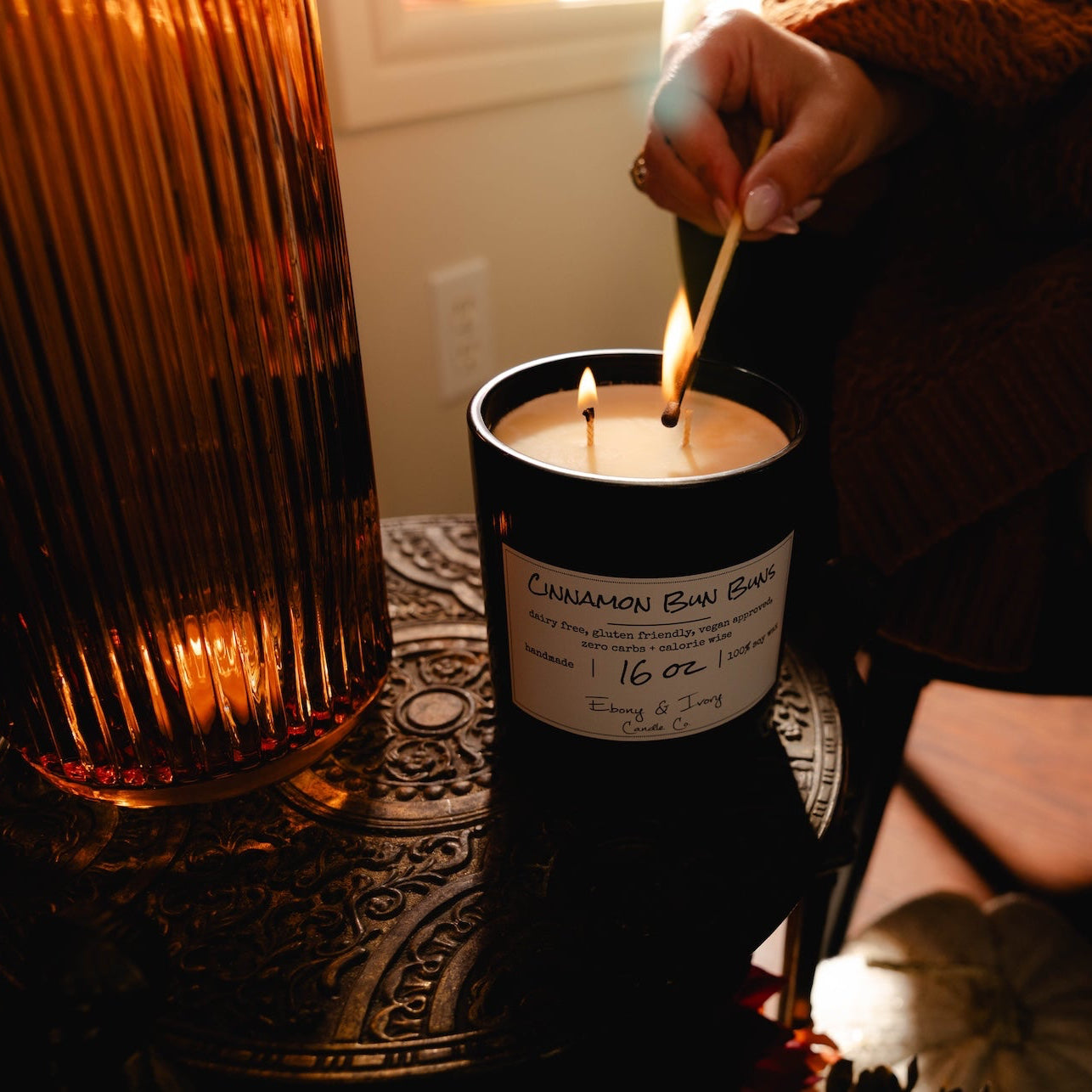 a black 16oz soy candle with a white lable named "cinnamon bun buns" on an iron garden table with autumn decor, being lit by a long match