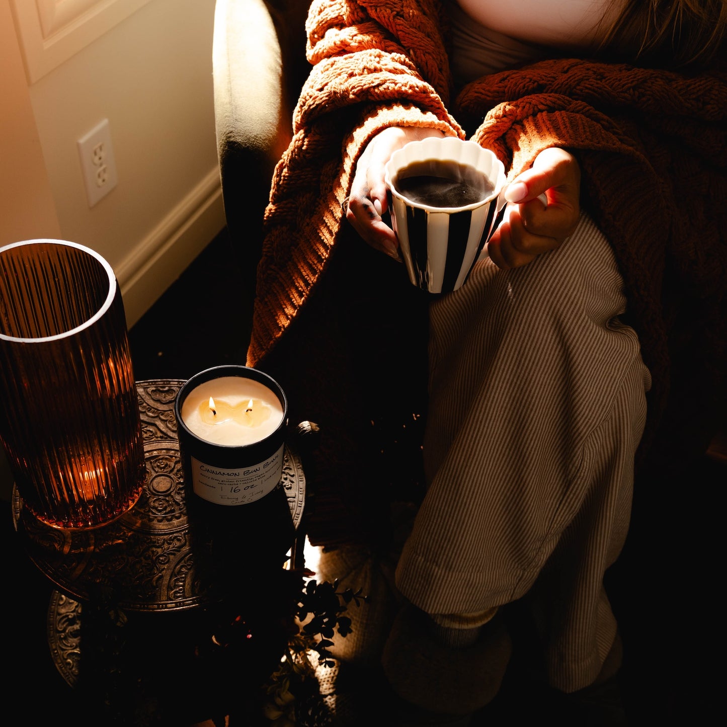 Black 16oz soy wax candle with a white label named "cinnamon bun buns" around autumn decor and a person in an fall sweater holding a striped mug of coffee