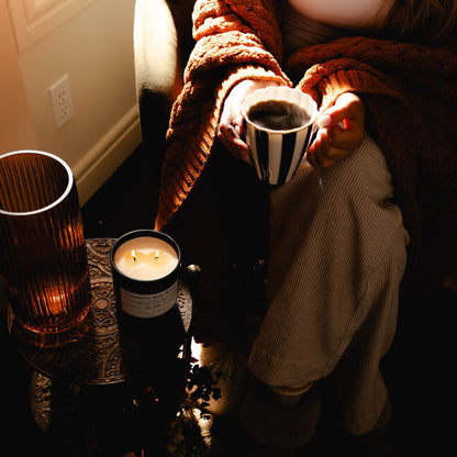 Black 16oz soy wax candle with a white label named "cinnamon bun buns" around autumn decor and a person in an fall sweater holding a striped mug of coffee
