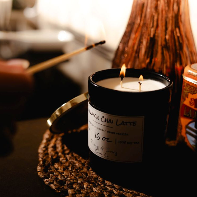 A black 16oz soy candle with two cotton wicks being lit by a match, sitting next to a tin of chai.