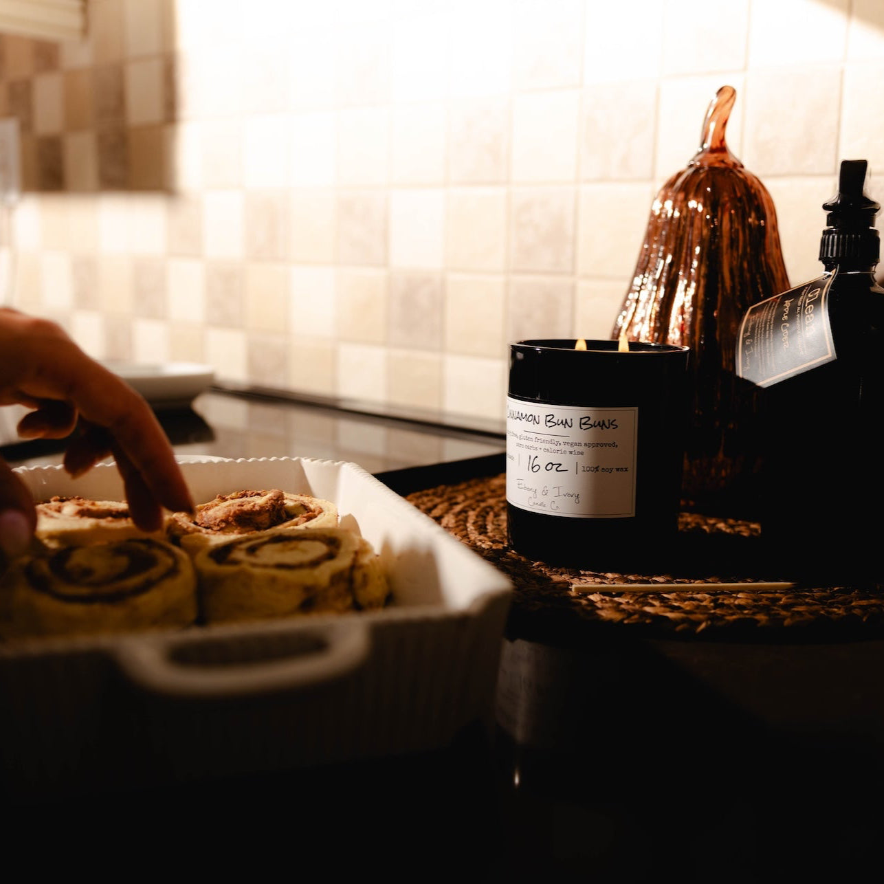 Person holding a tray of cinnamon buns with a candle and decorative bottle in the background. Cozy kitchen decor.