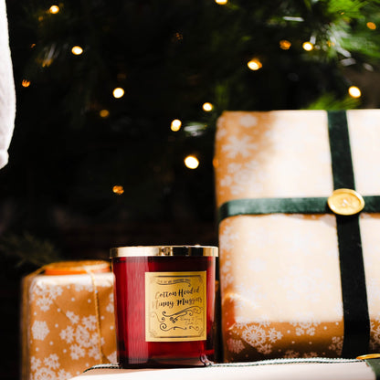 Red 16oz soy wax candle with a gold lid and a old label named "cotton headed ninny muggins" sitting under the christmas tree with wrapped presents. 