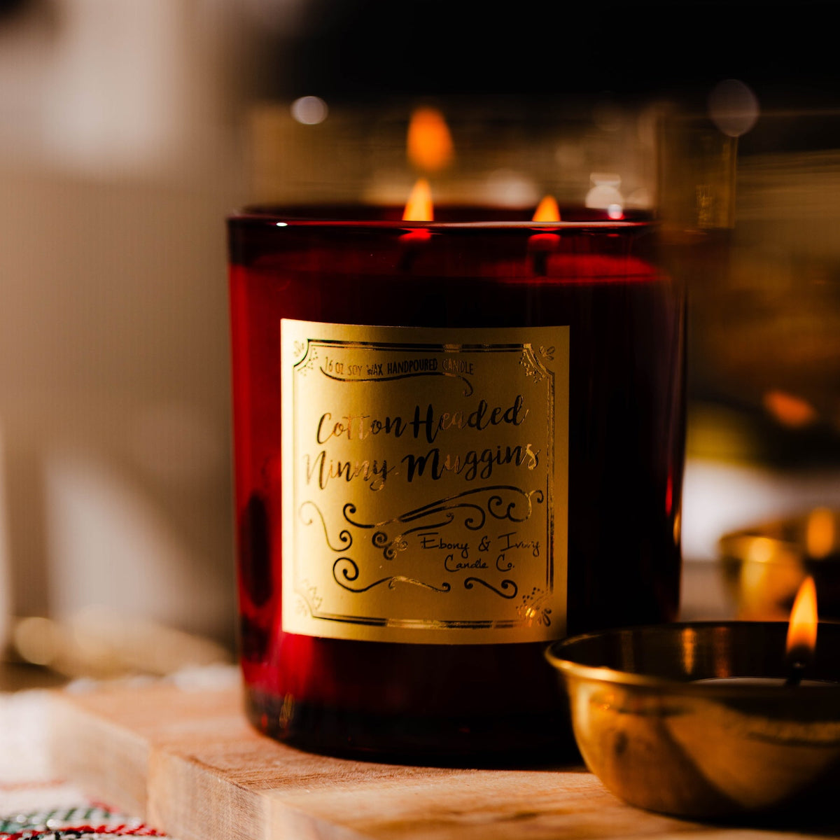 A red 16oz soy candle with a gold label named "cotton headed ninny muggins" sitting on a christmas dining table with tea lights in decorative gold holders