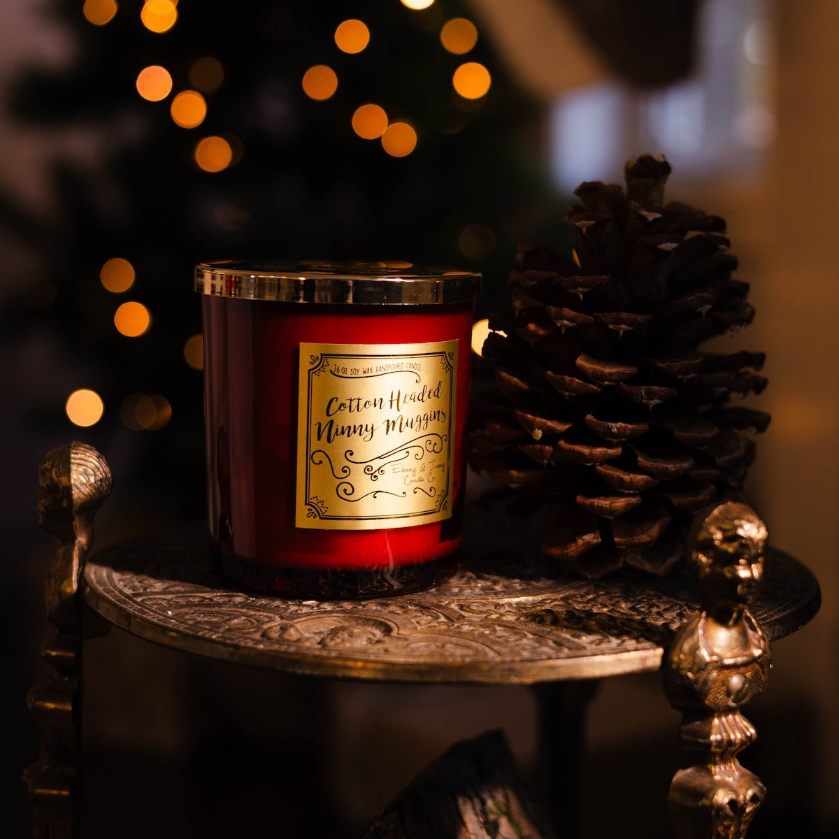 A red 16oz soy christmas candle with a gold lid and a gold label named "cotton headed ninny muggins" sitting on a gold metal side table with a pine cone and christmas lights.
