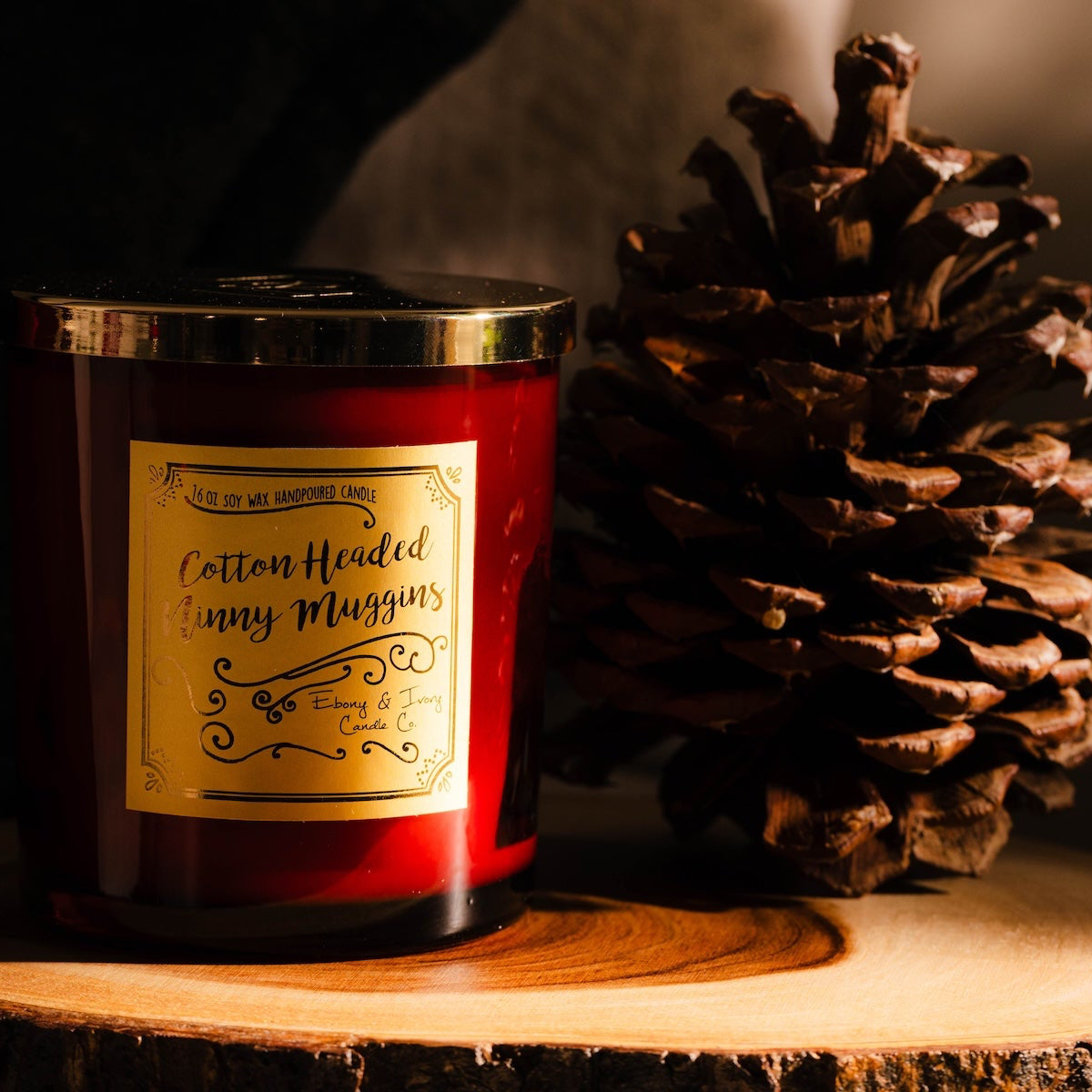 A red 16oz soy christmas candle with a gold lid and a gold label named "cotton headed ninny muggins"sitting on a wood surface with a pinecone.