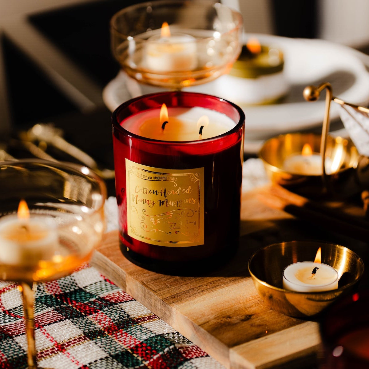 A red 16oz soy christmas candle with a gold label named "cotton headed ninny muggins" sitting on a christmas dining table.