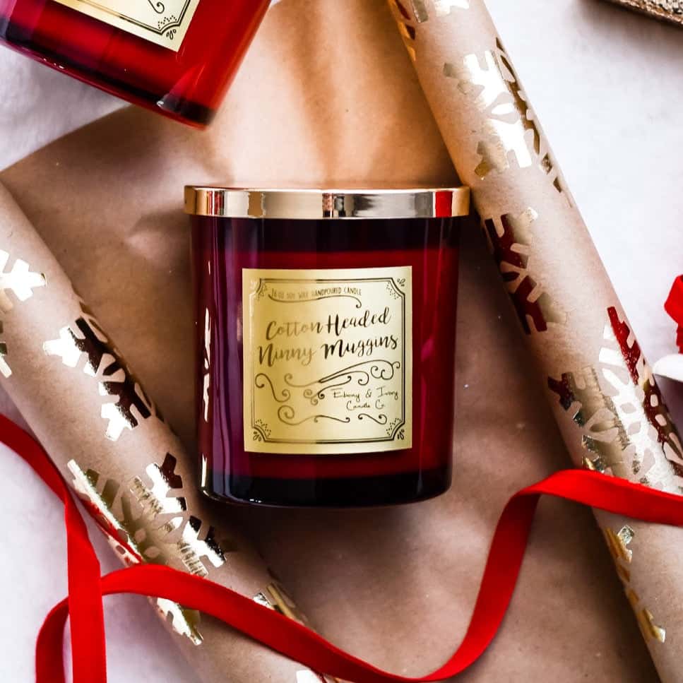 A red 16oz soy christmas candle with a gold lid and a gold label named "cotton headed ninny muggins" laying on christmas wrapping paper and surrounded by red velvet ribbon.