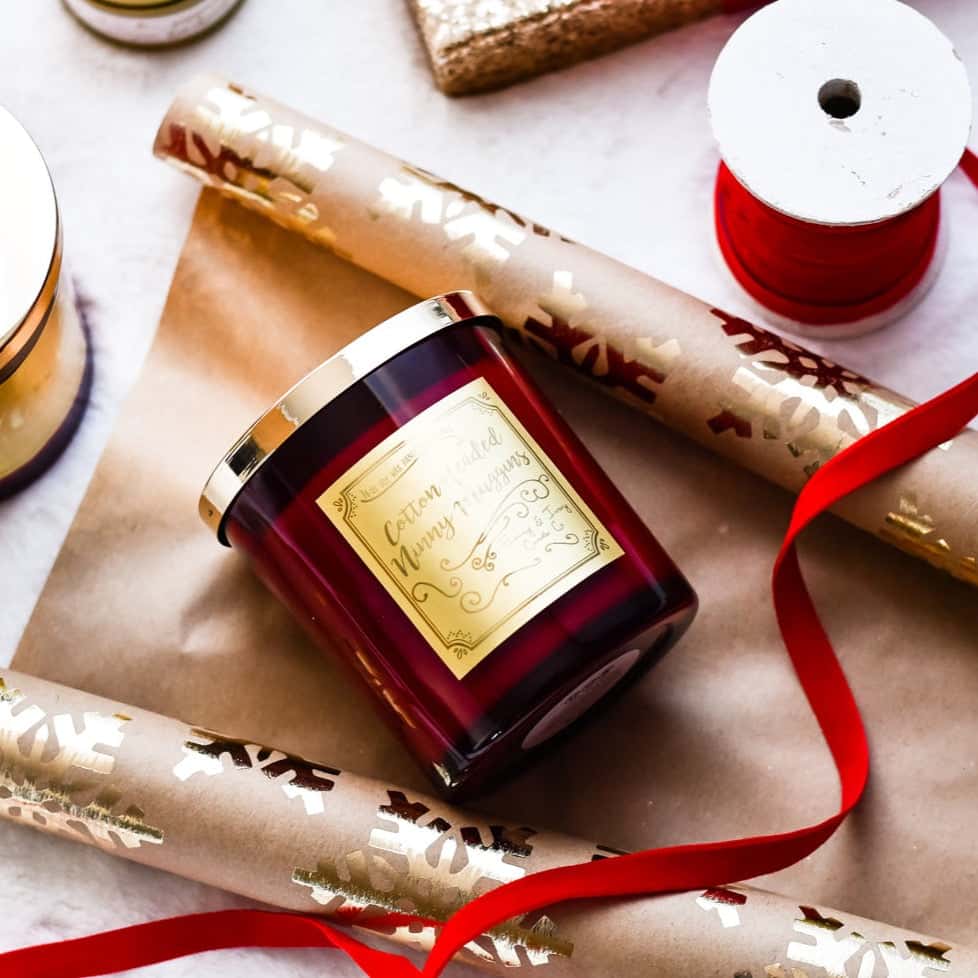 A red 16oz soy christmas candle with a gold lid and a gold label named "cotton headed ninny muggins" laying on brown and gold snowflake wrapping paper with red velvet ribbon.