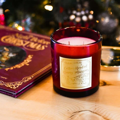a red 16oz soy christmas candle with a gold label named "cotton headed ninny muggins" sitting on a table with the book "the night before christmas"
