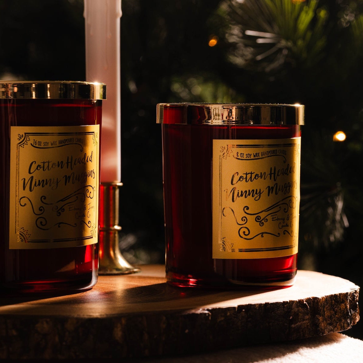 two red christmas soy candles with gold lids and gold labels named "cotton headed ninny muggins" sitting on a wood surface with christmas decorations. 