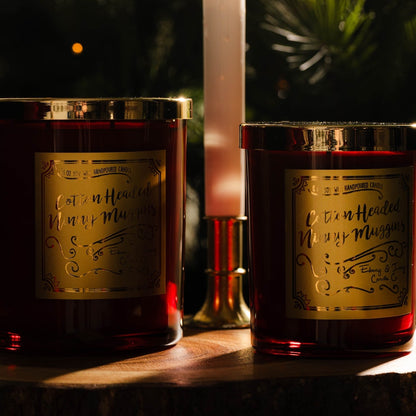 Two red christmas soy candles with gold lids and gold labels named "cotton headed ninny muggins" sitting on a table with christmas decor.