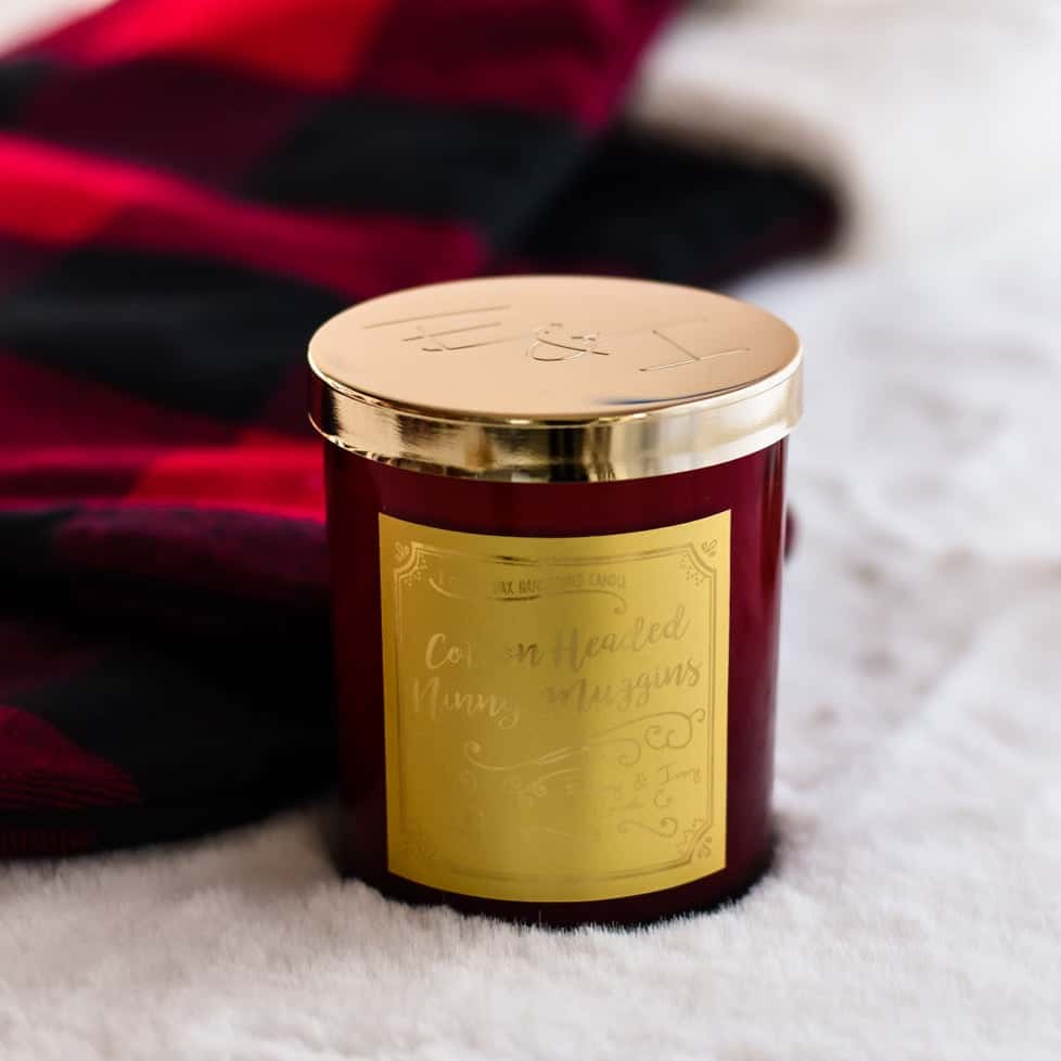 a red 8oz soy christmas candle with a gold lid and a gold label named "cotton headed ninny muggins" sitting on a fluffy white rug with a plaid christmas stocking.