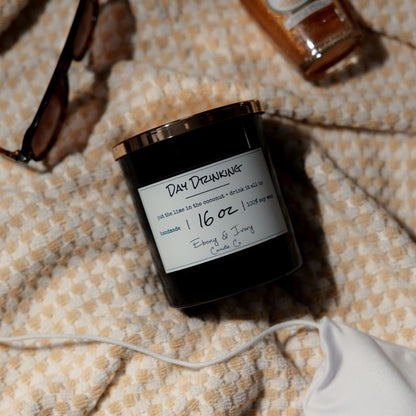 A black 16oz soy candle with a gold lid, labelled Day Drinking, laying on a beach towel. Made in lethbridge Alberta Canada