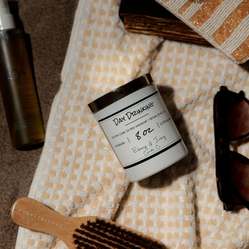 A white 8oz soy candle with a gold lid, labelled Day Drinking, laying on a beach towel. Made in Lethbridge Alberta Canada