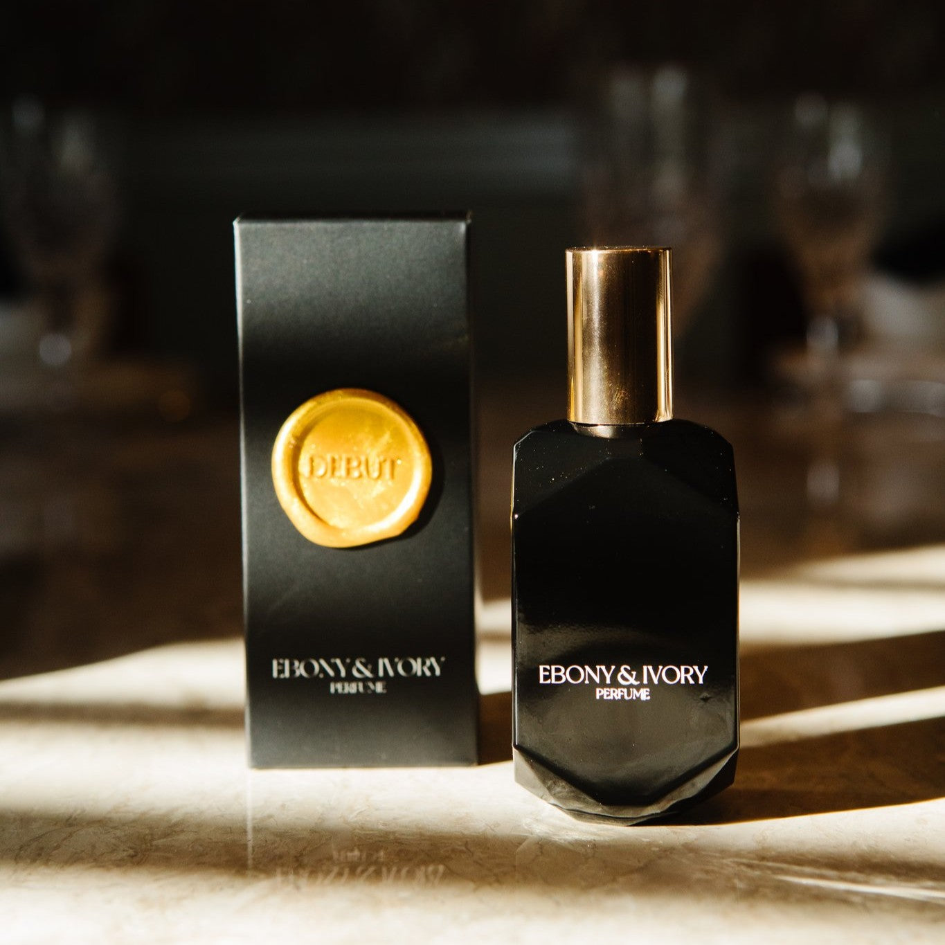a black geometric perfume bottle made by ebony & ivory candle co in Lethbridge alberta canada, sitting next to it's black box with a gold wax seal label named Debut, on a gold counter top with sun shining on them
