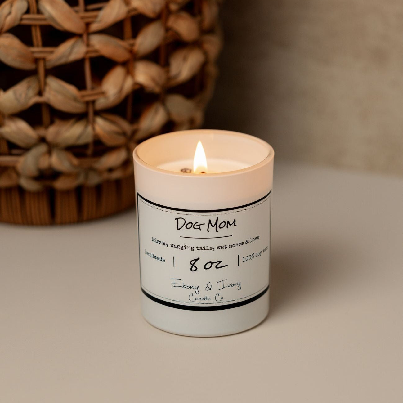 A white 8oz soy candle named Dog Mom sitting on a table with a wicker basket behind it. Made in Lethbridge Alberta Canada