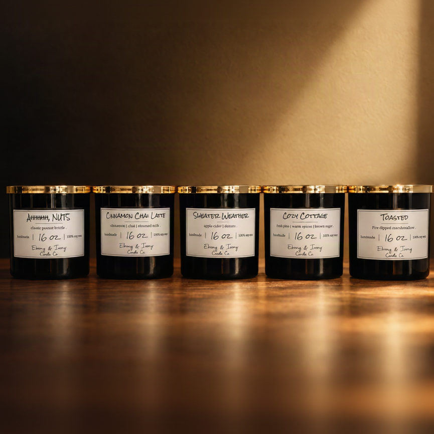 A set of 5 black 16oz soy candles with gold lids and white labels, from left to right they're named "Ahhhhh, NUTS" "Cinnamon Chai Latte" "Sweater Weather" "Cozy Cottage" "Toasted" On a brown tabled with warm autumn lighting