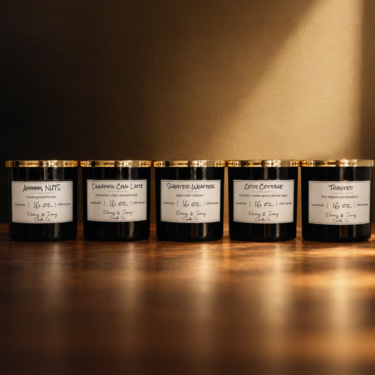 A set of 5 black 16oz soy candles with gold lids and white labels, from left to right they're named "Ahhhhh, NUTS" "Cinnamon Chai Latte" "Sweater Weather" "Cozy Cottage" "Toasted" On a brown tabled with warm autumn lighting