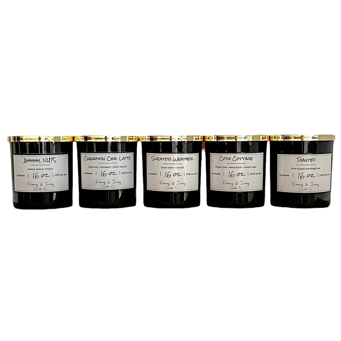 A set of 5 black 16oz soy candles with gold lids and white labels, from left to right they're named "Ahhhhh, NUTS" "Cinnamon Chai Latte" "Sweater Weather" "Cozy Cottage" "Toasted" 