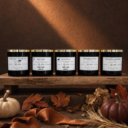 Five scented candles in black jars with gold lids and white labels are arranged on a wooden shelf in a rustic autumn setting with pumpkins, wheat, leaves, and a warm brown wall with a diagonal light beam.