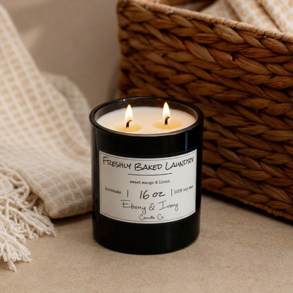 A black 16oz soy candle with a white label named "Freshly Baked Laundry" sitting on a counter with a white towel and a wicker basket. 