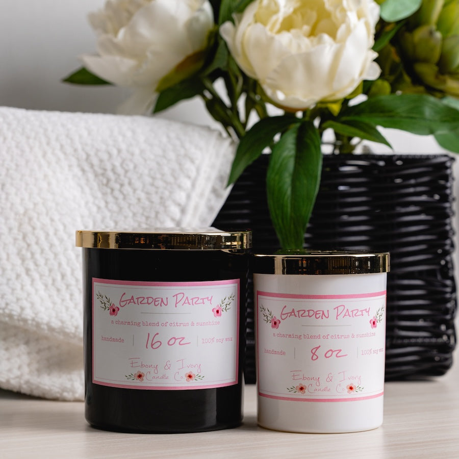 Two candles with labels on a surface, accompanied by a vase of white flowers. best soy candle Lethbridge Alberta canada. black and white candles with gold lid. 