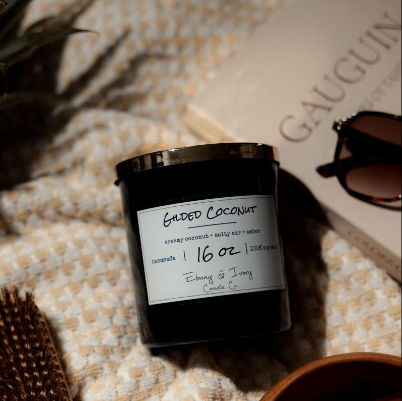A black 16oz soy candle with a gold lid, labelled 'Gilded Coconut,' Laying on a beach towel with a book and a pineapple. Made in Lethbridge Alberta Canada