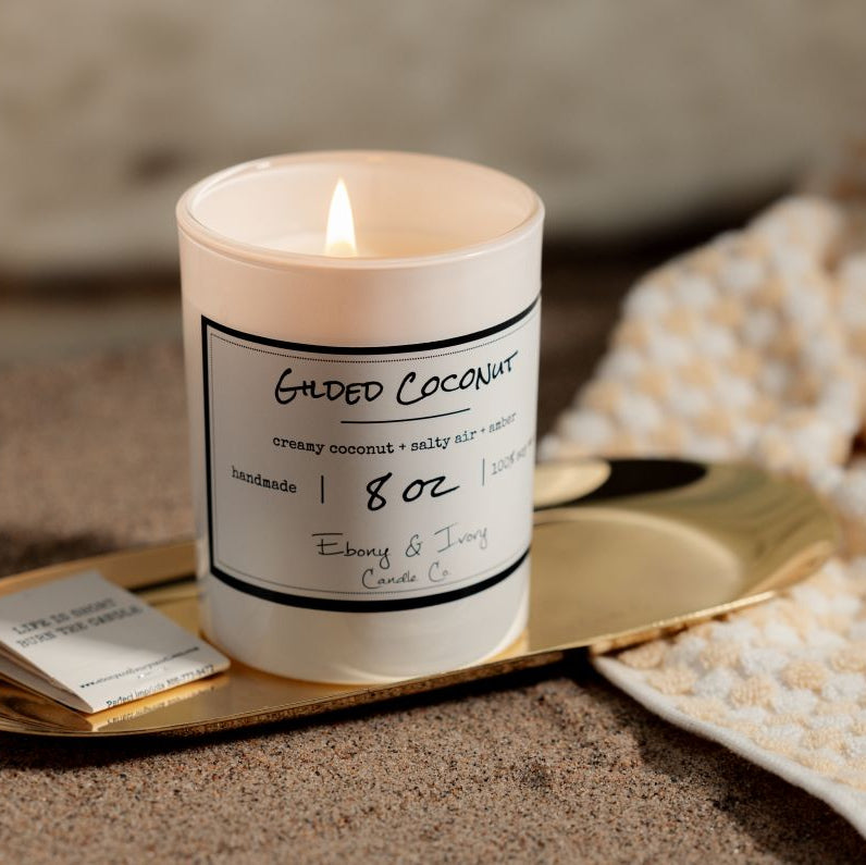 A white 8oz soy candle labelled 'Gilded Coconut' on the beach on a gold tray with a book of matches and a beach towel. 