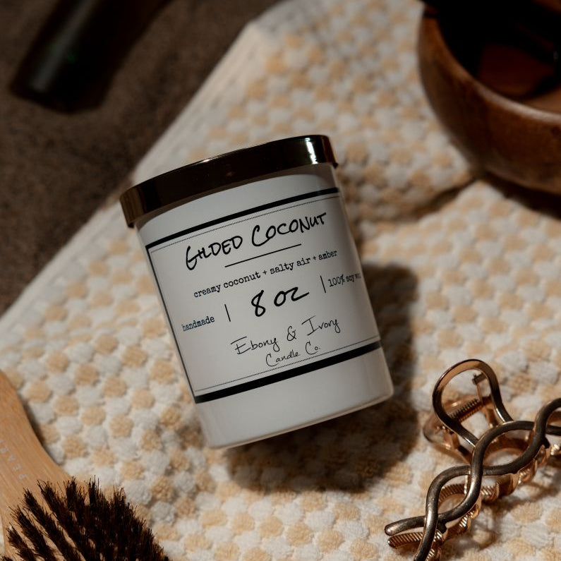 A white 8oz candle with a gold lid, labelled 'Gilded Coconut', laying on a beach towel. Made in Lethbridge Alberta Canada