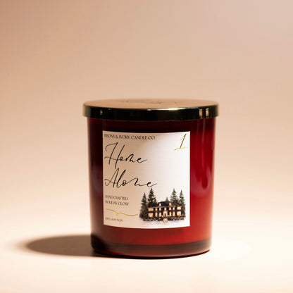 A red 16oz soy christmas candle with a gold lid and a white label named "home alone 1" on a beige background