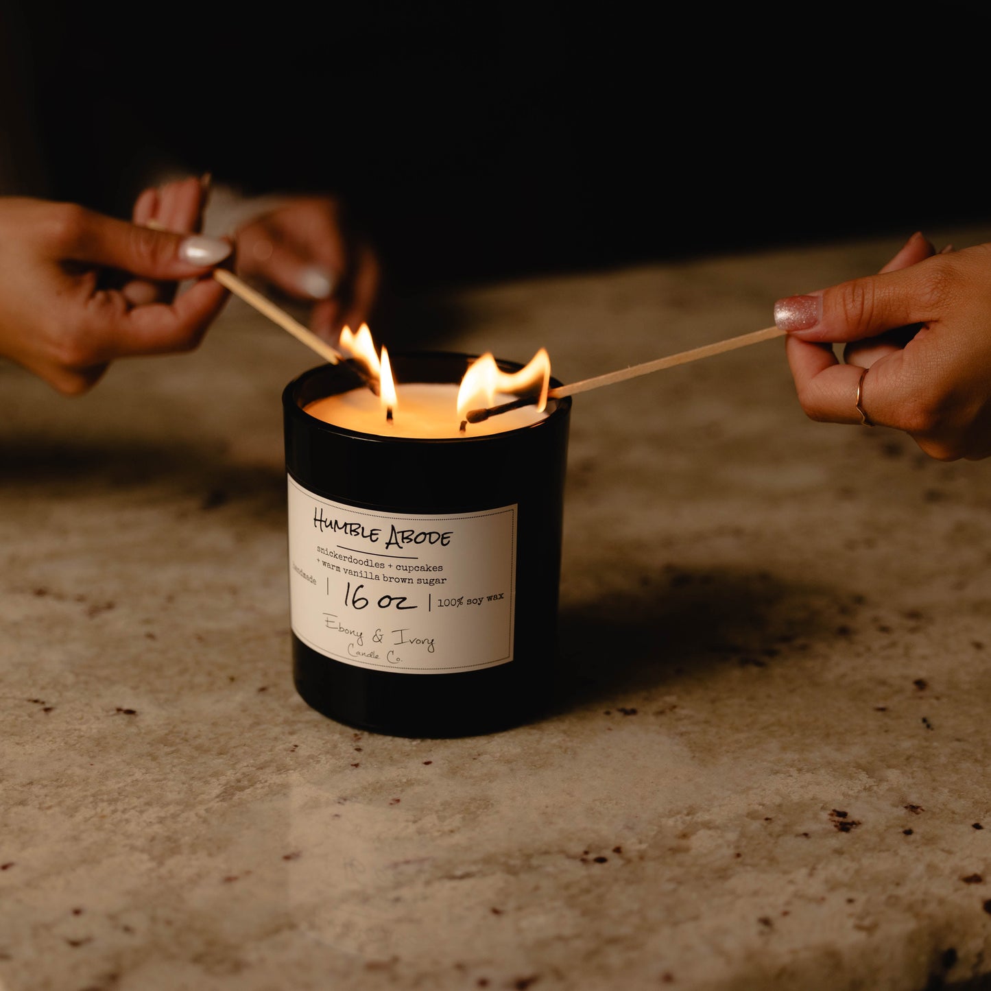 A black 16oz jar soy candle with a gold lid and a white label named Humble Abode, on a granite counter top with two people lighting the wicks with match sticks, made by Ebony & Ivory Candle Co in Lethbridge, Alberta, Canada
