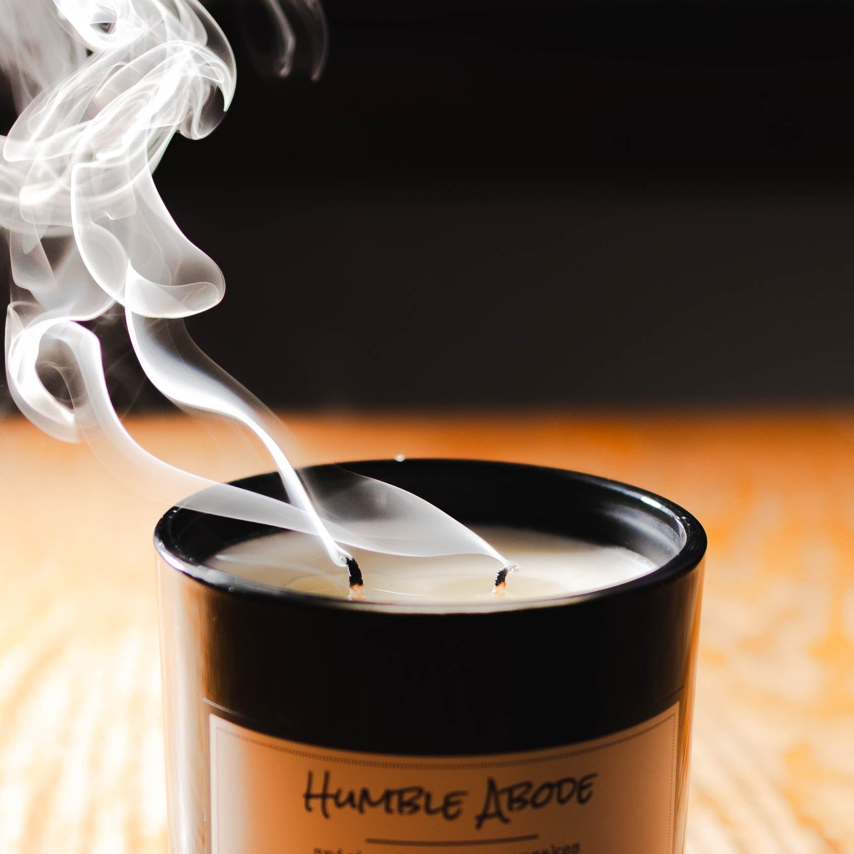 A black 16oz jar soy candle with a gold lid and a white label named Humble Abode, with the wicks smoking after just being blown out, made by Ebony & Ivory Candle Co in Lethbridge, Alberta, Canada