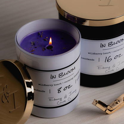 Two candles sitting next to eachother. The first one is a white 8oz jar soy candle with a gold lid sitting next to it, the wick is lit and it show cases purple coloured wax with wildflowers on top, it has a white label named In Bloom. The second candle is a black 16oz jar soy candle with a gold lid on top and a white label named In Bloom, they're sitting on a wood surface with gold wick trimmers nearby. They're made by Ebony & Ivory Candle Co in Lethbridge, Alberta, Canada