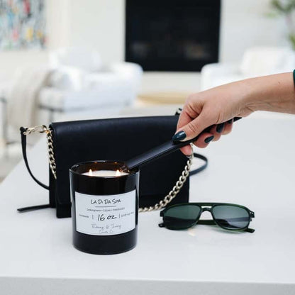 A black jar 16oz soy candle with a white label named La Di Da Spa, on a white table, being lit by an ebony & ivory candle co electric usb lighter, with a black purse behind it, made by Ebony & Ivory Candle Co in Lethbridge, Alberta, Canada