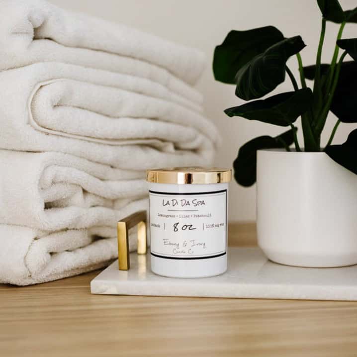 A white jar 8oz soy candle with a gold lid and a white label named La Di Da Spa, on a white tray with a potted plant and towels behind it, made by Ebony & Ivory Candle Co in Lethbridge, Alberta, Canada