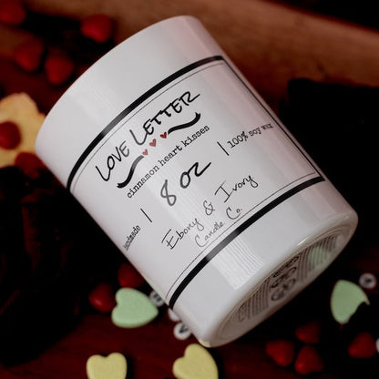 White candle jar with black text on a wooden surface with heart-shaped objects. cinnamon heart candy soy candle Canada for valentines day.