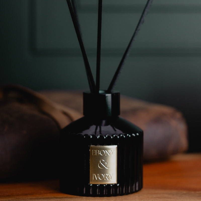 A black 100ml ribbed luxx reed diffuser with black diffuser reeds and a gold Ebony & Ivory sticker, sitting on a wooden table with a leather journal behind it, made by Ebony & Ivory Candle Co in Lethbridge, Alberta, Canada