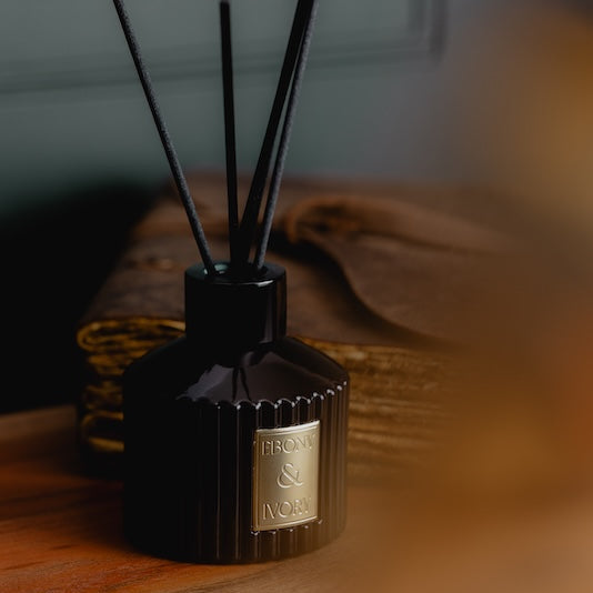 A black 100ml ribbed luxx reed diffuser with black diffuser reeds and a gold Ebony & Ivory sticker, sitting on a wooden table with a leather journal behind it and blurring infront, made by Ebony & Ivory Candle Co in Lethbridge, Alberta, Canada
