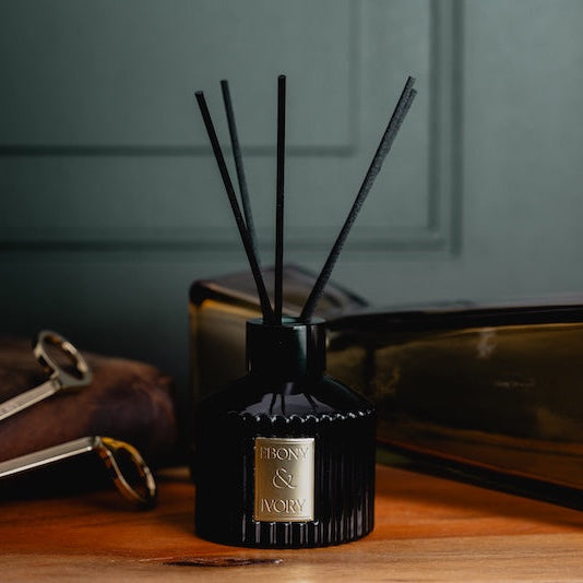 A black 100ml ribbed luxx reed diffuser with black diffuser reeds and a gold Ebony & Ivory sticker, sitting on a wooden table with a leather journal and gold candle wick trimmers behind it, made by Ebony & Ivory Candle Co in Lethbridge, Alberta, Canada