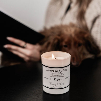 A white jar 8oz soy candle with a gold lid and a white label named Mom's in a Mood, on a black table, with a person reading a book and a dog in their lap behind it, made by Ebony & Ivory Candle Co in Lethbridge, Alberta, Canada
