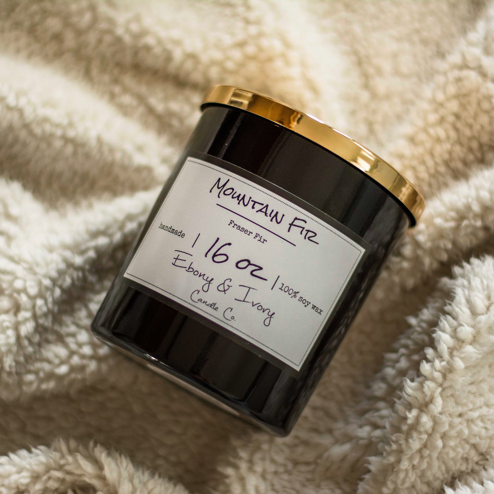 A black jar 16oz soy candle with a gold lid and a white label named Mountain Fir, on a fluffy white cozy blanket, made by Ebony & Ivory Candle Co in Lethbridge, Alberta, Canada