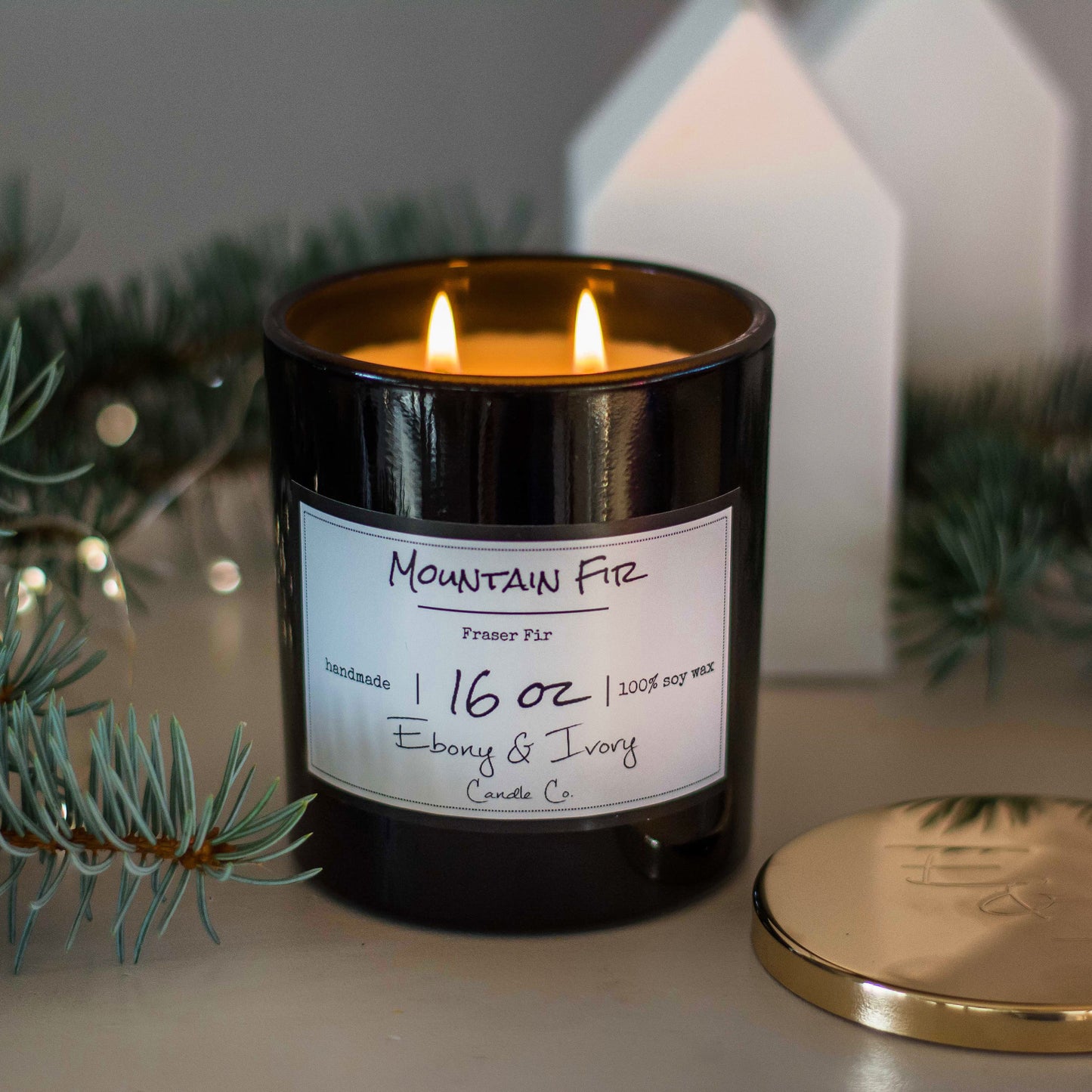 A black jar 16oz soy candle with a gold lid and a white label named Mountain Fir, on a table with fraser fir needles and branches around it, made by Ebony & Ivory Candle Co in Lethbridge, Alberta, Canada