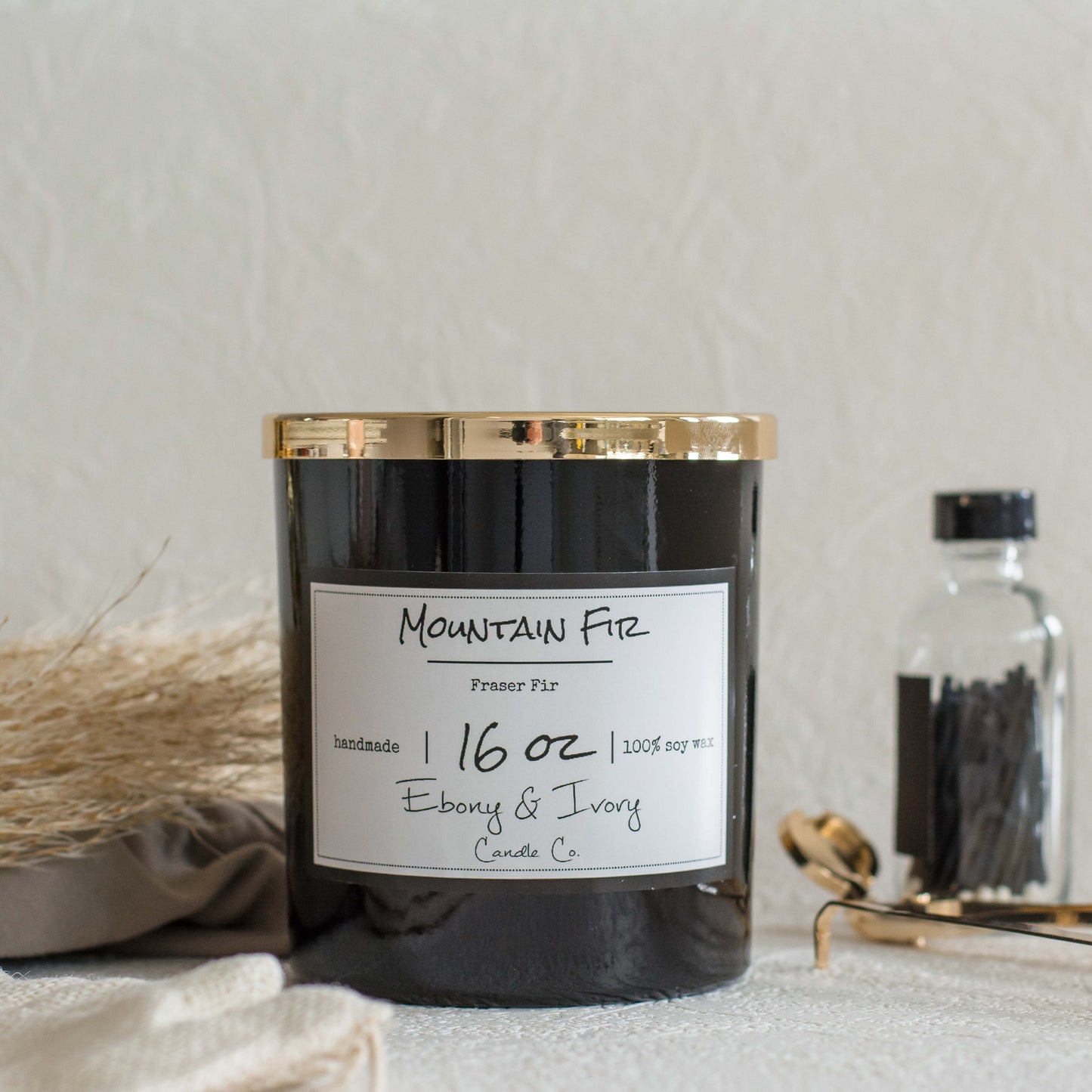 A black jar 16oz soy candle with a gold lid and a white label named Mountain Fir, on a white table with gold trimmers and a bottle of matches behind it, made by Ebony & Ivory Candle Co in Lethbridge, Alberta, Canada