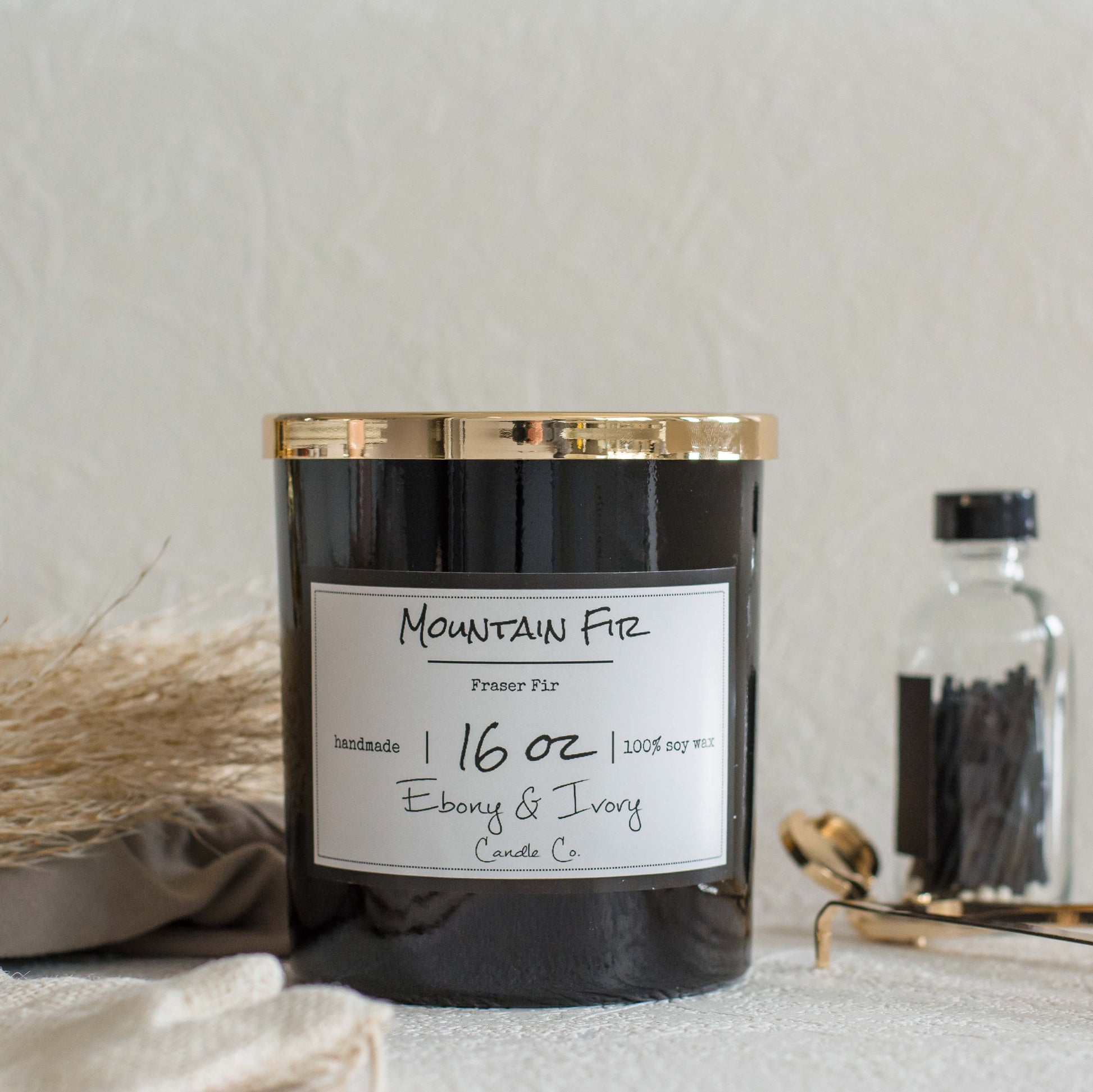 A black jar 16oz soy candle with a gold lid and a white label named Mountain Fir, on a white table with gold trimmers and a bottle of matches behind it, made by Ebony & Ivory Candle Co in Lethbridge, Alberta, Canada