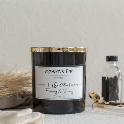 A black jar 16oz soy candle with a gold lid and a white label named Mountain Fir, on a white table with gold trimmers and a bottle of matches behind it, made by Ebony & Ivory Candle Co in Lethbridge, Alberta, Canada