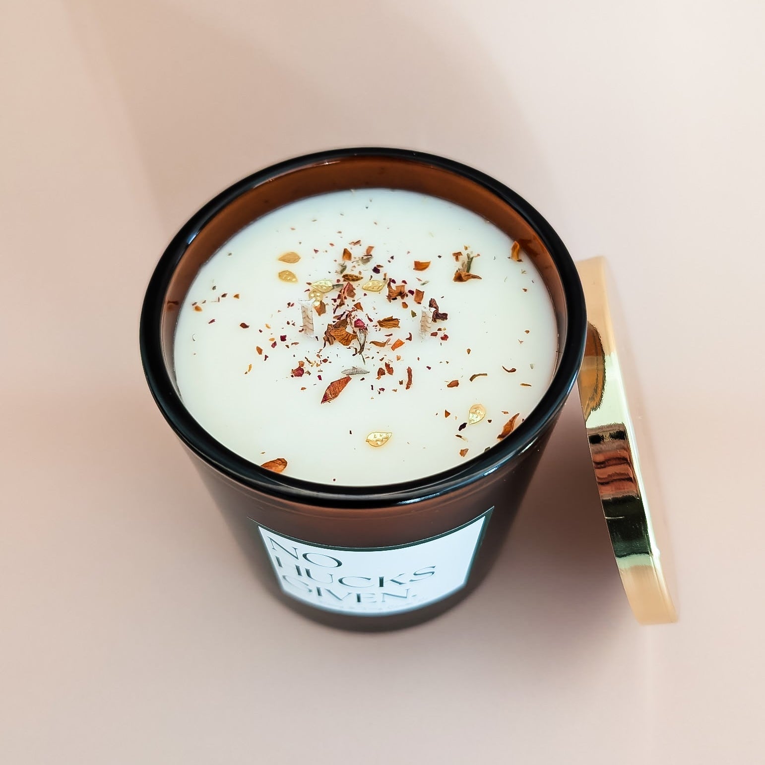 A top down view of an amber jar 16oz soy candle with dried rose petals and gold leaf glitters, a gold lid and a white label named No Hucks Given, on an ivory background, made by Ebony & Ivory Candle Co in Lethbridge, Alberta, Canada