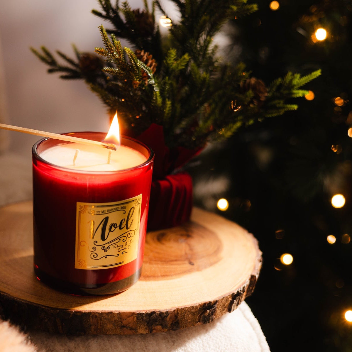 A red 16oz soy christmas candle with a gold lid named "noel" being lit by a match infront of a christmas tree with lights.