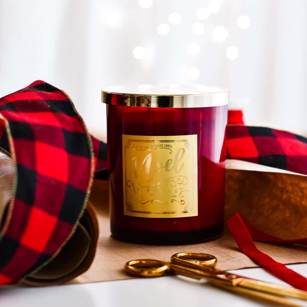 a red 16oz soy christmas candle with a gold lid and a gold label named "noel" sitting on wrapping paper and surrounded by plaid ribbon.