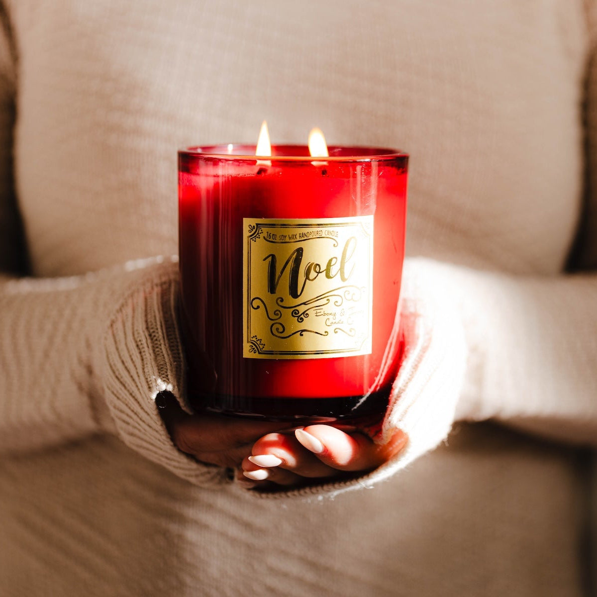 A red 16oz soy christmas candle with a gold label named "noel" being held by a person wearing a beige sweater.